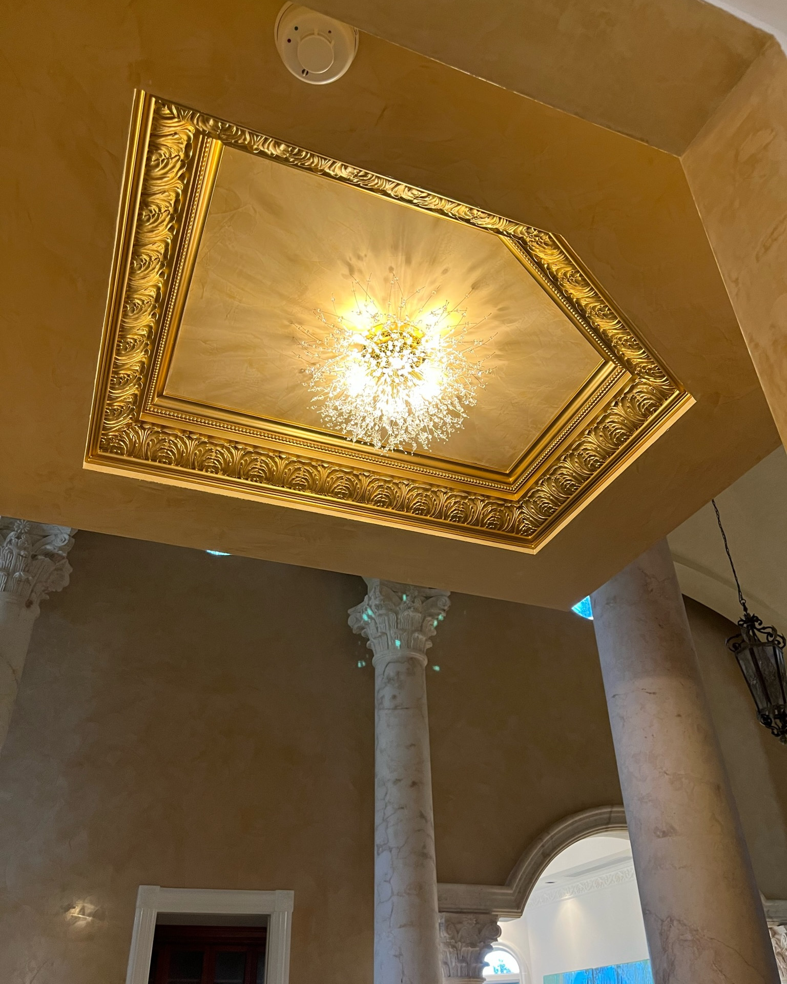 Gilded coffered ceiling panel framed by ornate gold acanthus molding with a starburst crystal light fixture, above marble columns with carved capitals.
