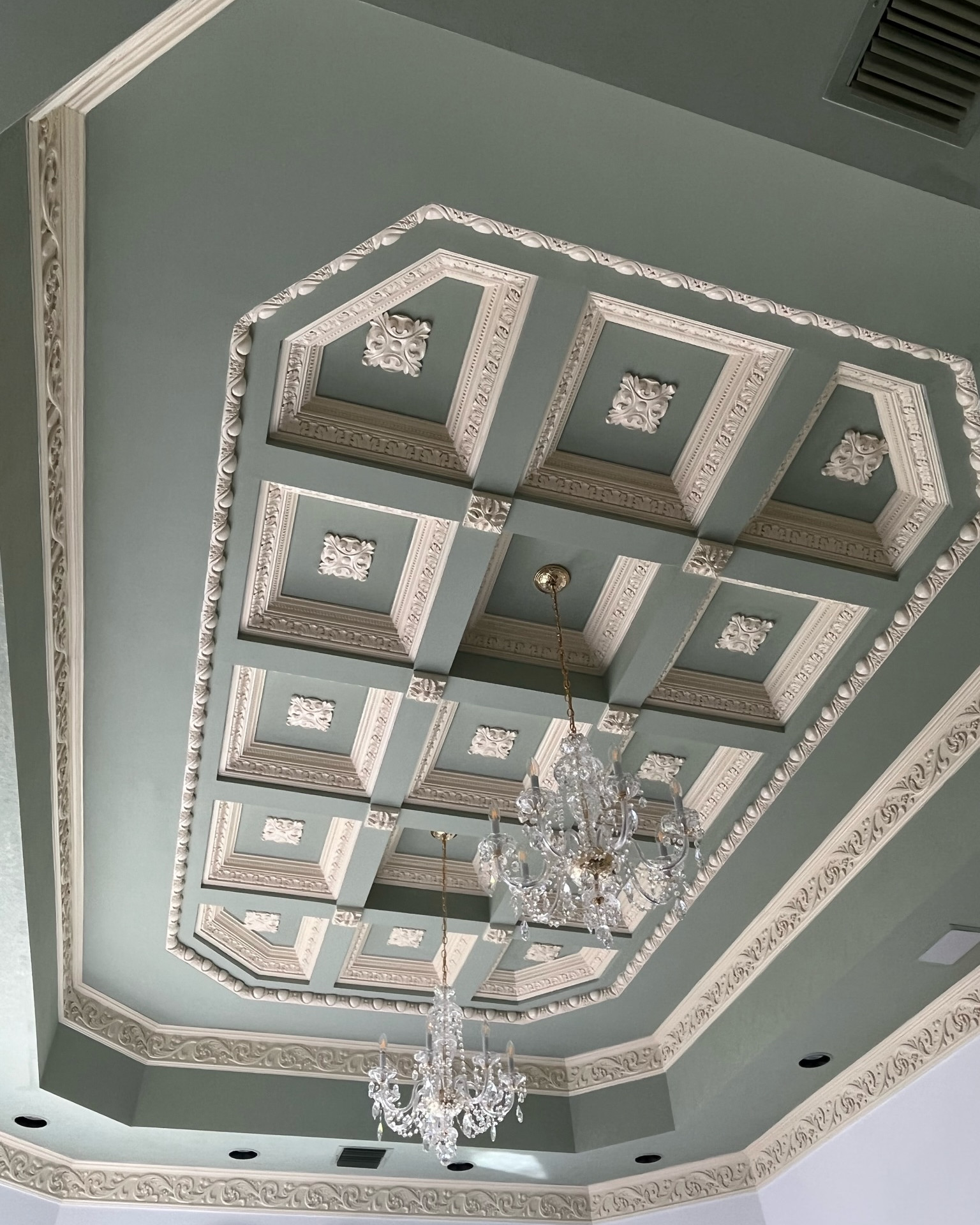 Sage green coffered ceiling with white plaster moldings, rosette medallions in each recessed panel and two crystal chandeliers hanging from gilt chains.