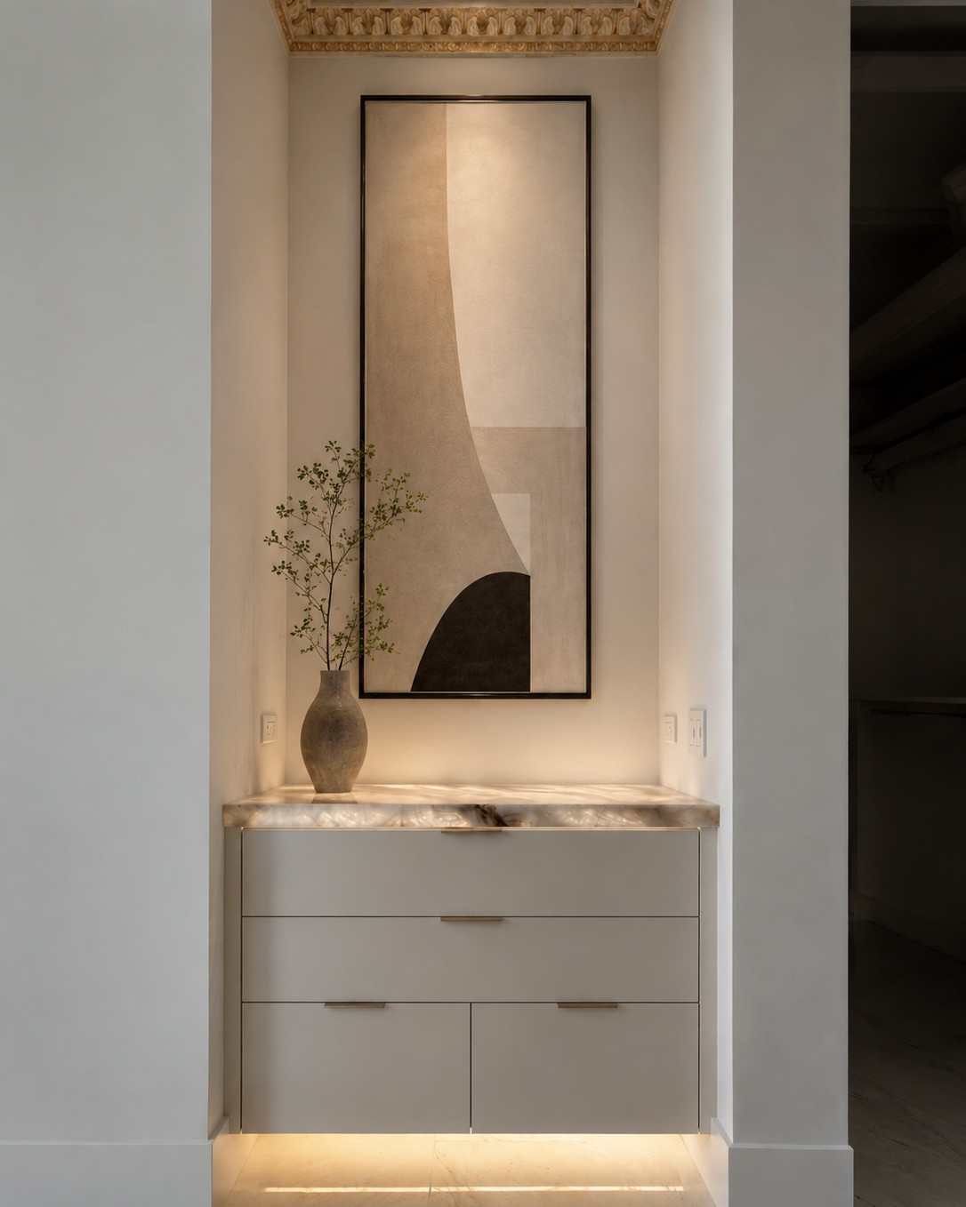 Recessed vestibule with a backlit onyx counter, pale grey millwork drawers with slim bar pulls, a tall framed abstract artwork, olive branches in a stoneware vase and a gilded cornice.
