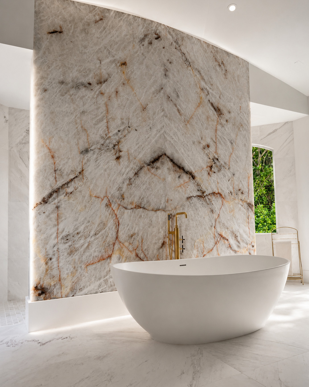 Freestanding white stone bathtub in front of a backlit bookmatched quartzite feature wall, with a brass floor-mounted tub filler and hand shower, arched window and marble floor.