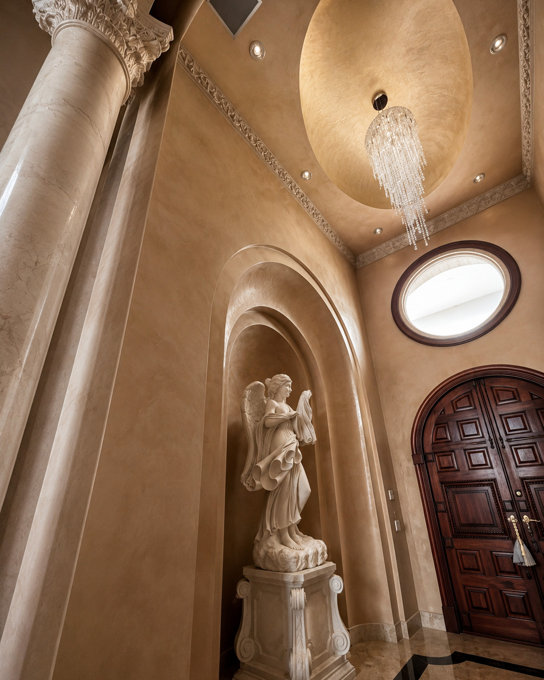 Double-height foyer with a domed ceiling, crystal waterfall chandelier, carved marble angel statue in an arched niche, oval window and dark wood arched double doors over a polished marble floor.