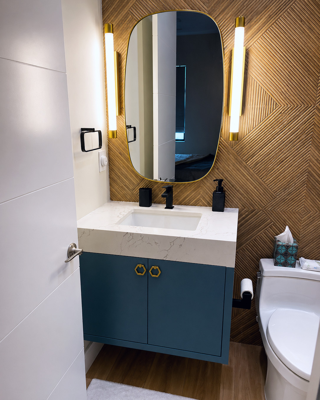 Powder room with a teal floating vanity, brass hexagonal pulls, a white quartz countertop with an undermount sink, an oval brass-framed mirror, brass linear sconces, and a textured wood-tone tile wall.