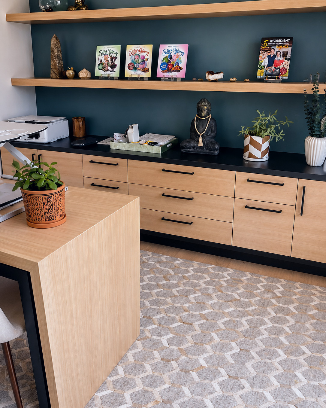 Detail of a home office credenza with white oak fronts, black bar pulls and a black countertop against a teal wall, with white oak floating shelves above and a white oak desk in the foreground.