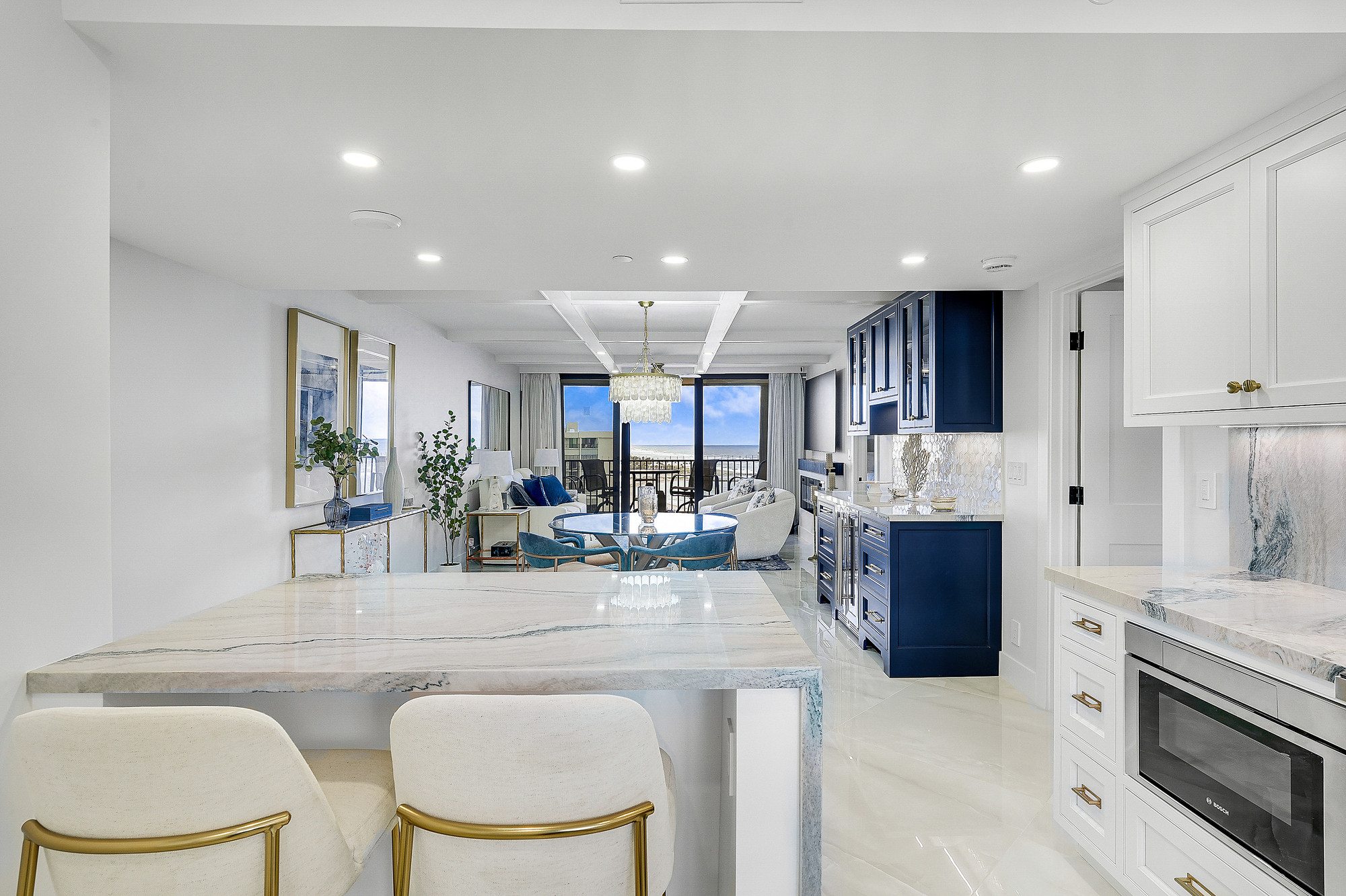 Open kitchen and living area with a veined quartzite peninsula and waterfall edge, upholstered bar stools with brass frames, navy blue cabinetry with glass-front uppers, coffered ceiling, capiz shell chandelier and sliding doors opening to an ocean view.
