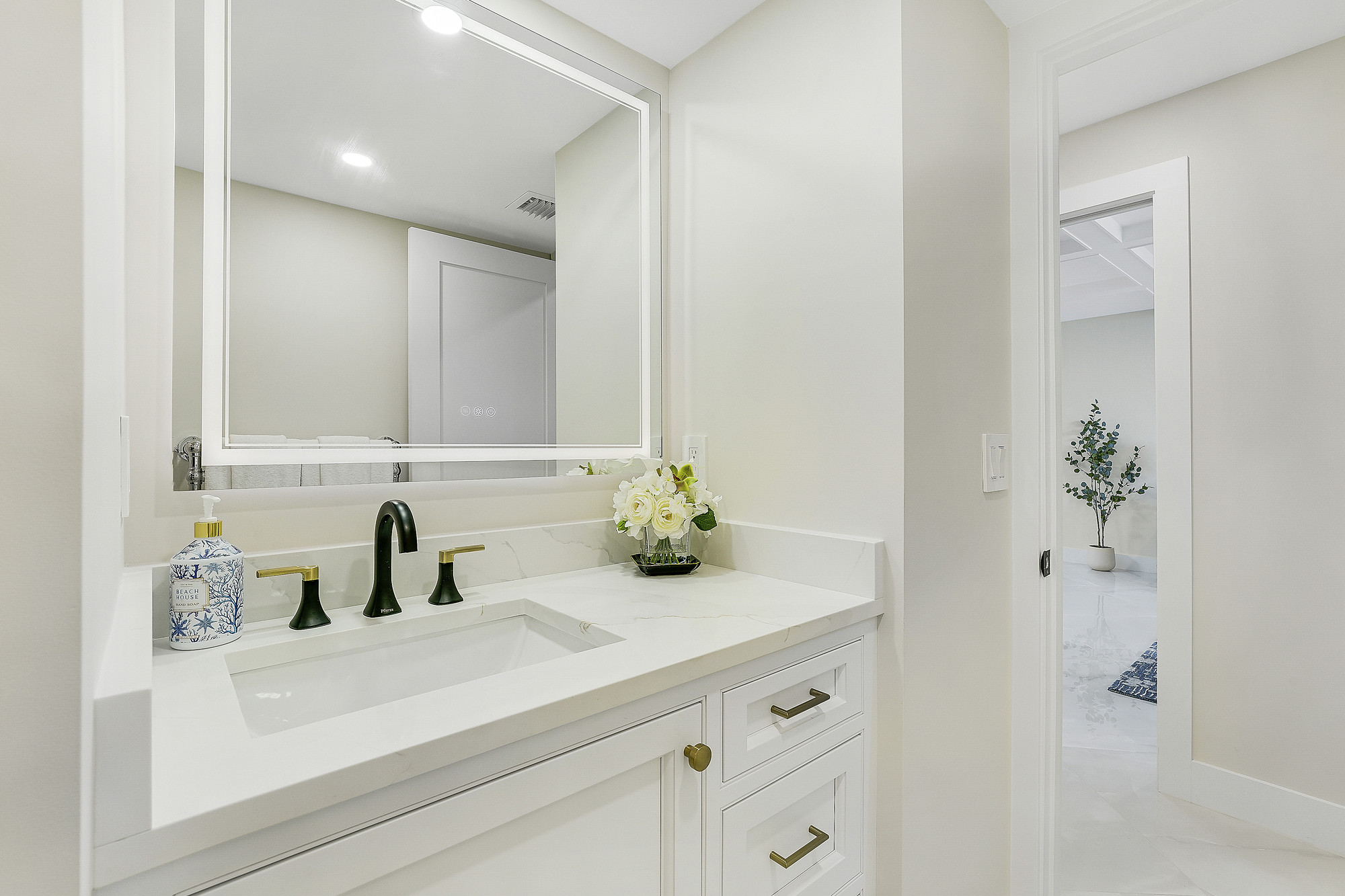 Powder room with a white quartz vanity top and integrated backsplash, black and brass widespread faucet, backlit rectangular mirror, white cabinetry with brass pulls and a doorway opening onto a polished marble-look floor.
