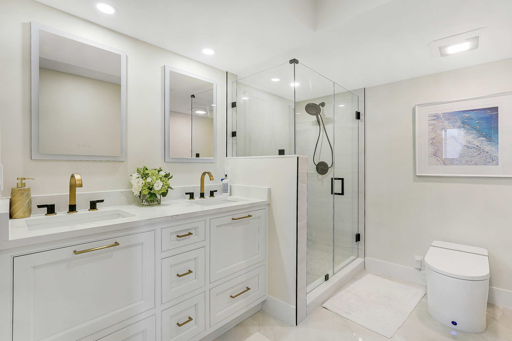 Primary bathroom with a double vanity in white inset cabinetry with brass pulls, white quartz countertop, two backlit mirrors, brass and matte black faucets, frameless glass corner shower with a half wall and a framed coastal artwork.