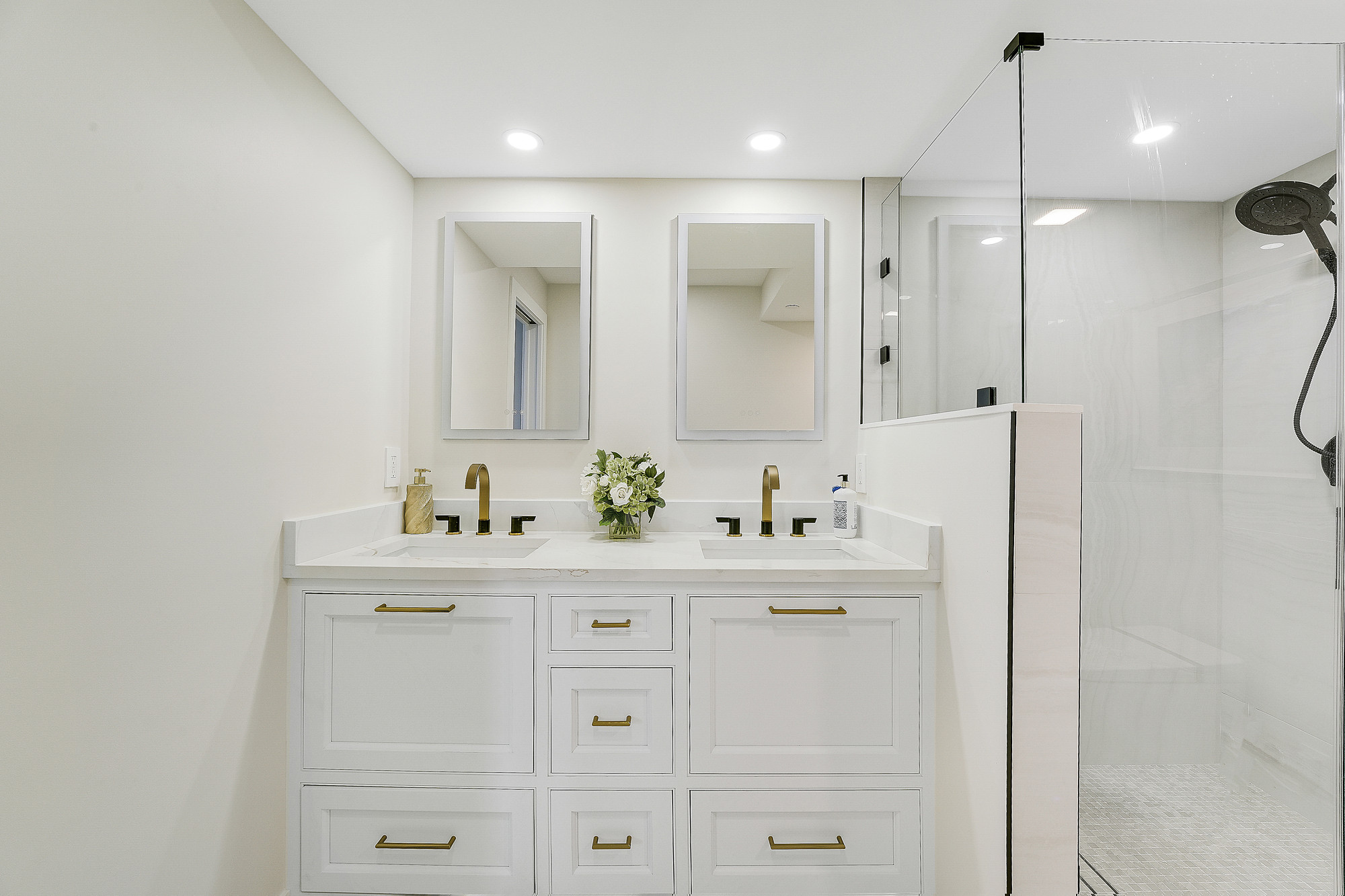 Frontal view of a double vanity in white inset cabinetry with brass pulls, white quartz countertop with two undermount sinks, brass and black faucets, two backlit mirrors and a frameless glass shower with a mosaic floor.