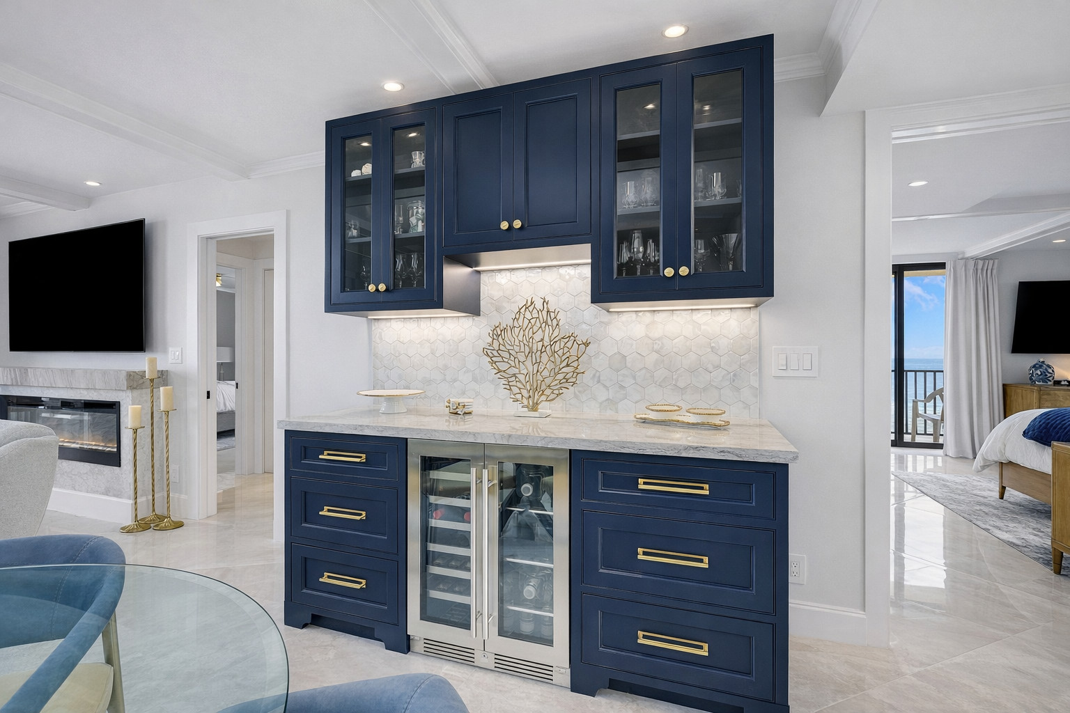 Navy blue inset bar cabinetry with brushed brass hardware, a marble countertop, pearl hexagon mosaic backsplash, glass-front upper cabinets and a stainless beverage cooler, opening to a bedroom with an ocean view