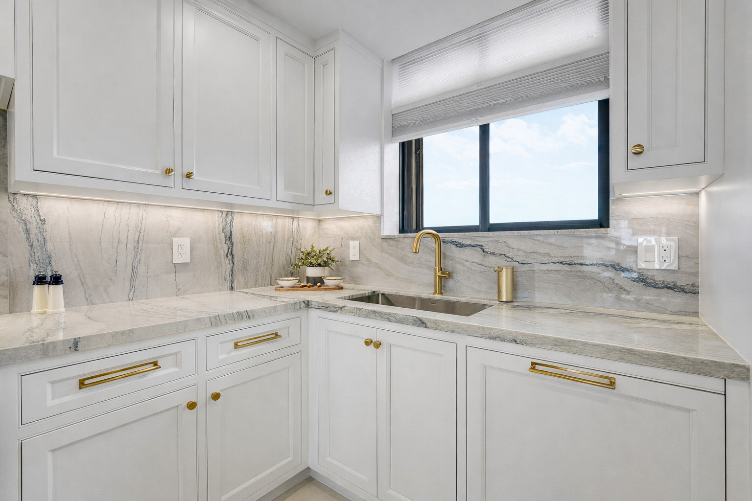 Corner of a white kitchen with a brushed brass gooseneck faucet, an undermount stainless sink, a full-height marble backsplash and inset cabinetry with brass pulls