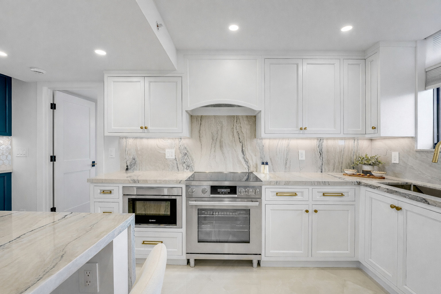White inset kitchen cabinetry with a book-matched marble slab backsplash, an arched hood over a stainless range, a built-in wall oven and brushed brass hardware