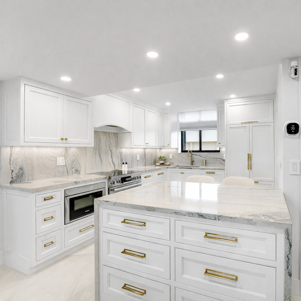 Wide view of a white kitchen with a marble-topped drawer island, a panel-ready refrigerator, a stainless range under an arched hood and a built-in wall oven
