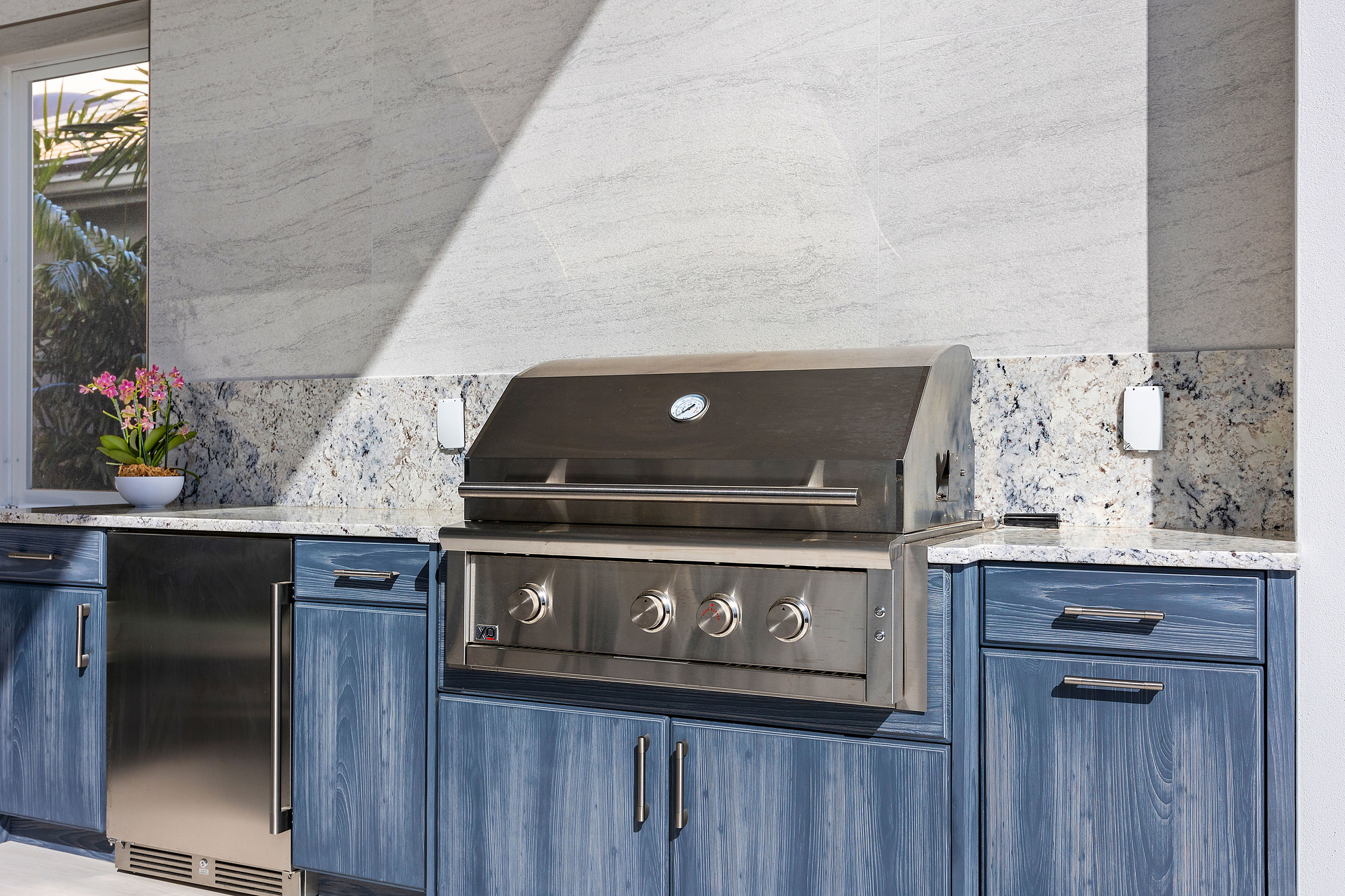 Outdoor kitchen with blue cerused wood cabinetry, speckled granite countertop and backsplash, a stainless steel built-in gas grill and an undercounter stainless refrigerator against a large-format stone slab wall.