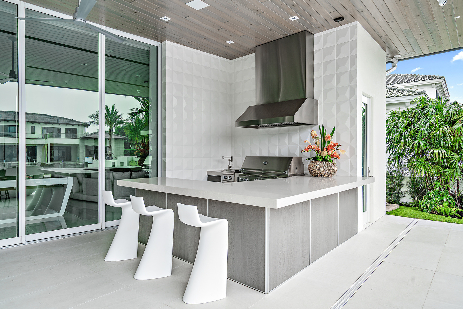 Covered terrace kitchen with a white quartz peninsula, three white sculptural bar stools, grey wood-grain cabinet fronts, a white three-dimensional tile wall, stainless steel hood and grill, framed by sliding glass doors and tropical planting.