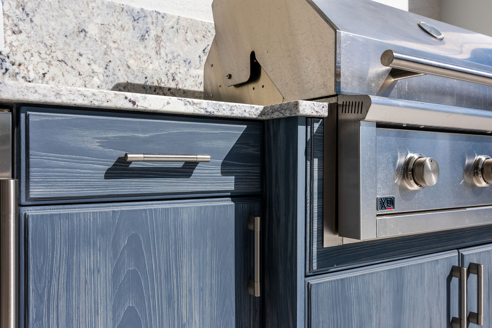 Close-up of blue cerused wood cabinet fronts with tubular stainless steel handles, a granite countertop edge and the control panel of a built-in stainless gas grill.