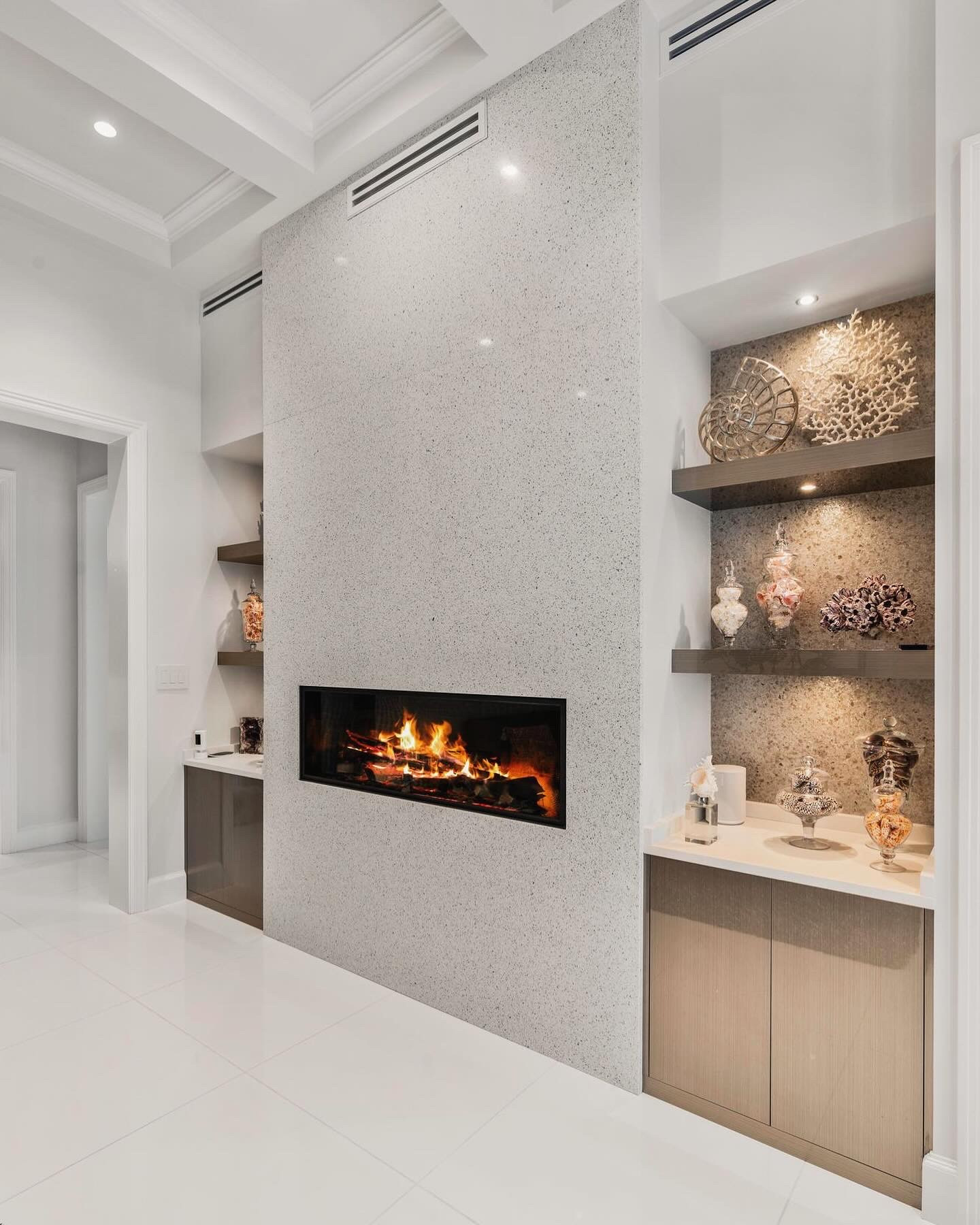 Floor-to-ceiling terrazzo fireplace wall with a recessed linear gas fireplace, flanked by lit niches with floating wood shelves holding coral, shells and glass apothecary jars, a stone counter and light oak base cabinets over polished white large-format tile.