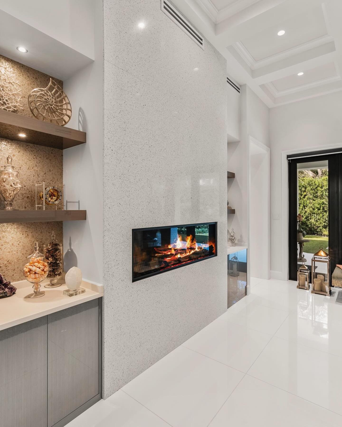 Terrazzo fireplace wall seen from the built-in bar side, with backlit niches holding shells, an amethyst geode and glass apothecary jars, high-gloss grey base cabinets with a stone countertop, a coffered ceiling and black-framed glass doors opening to a green garden.