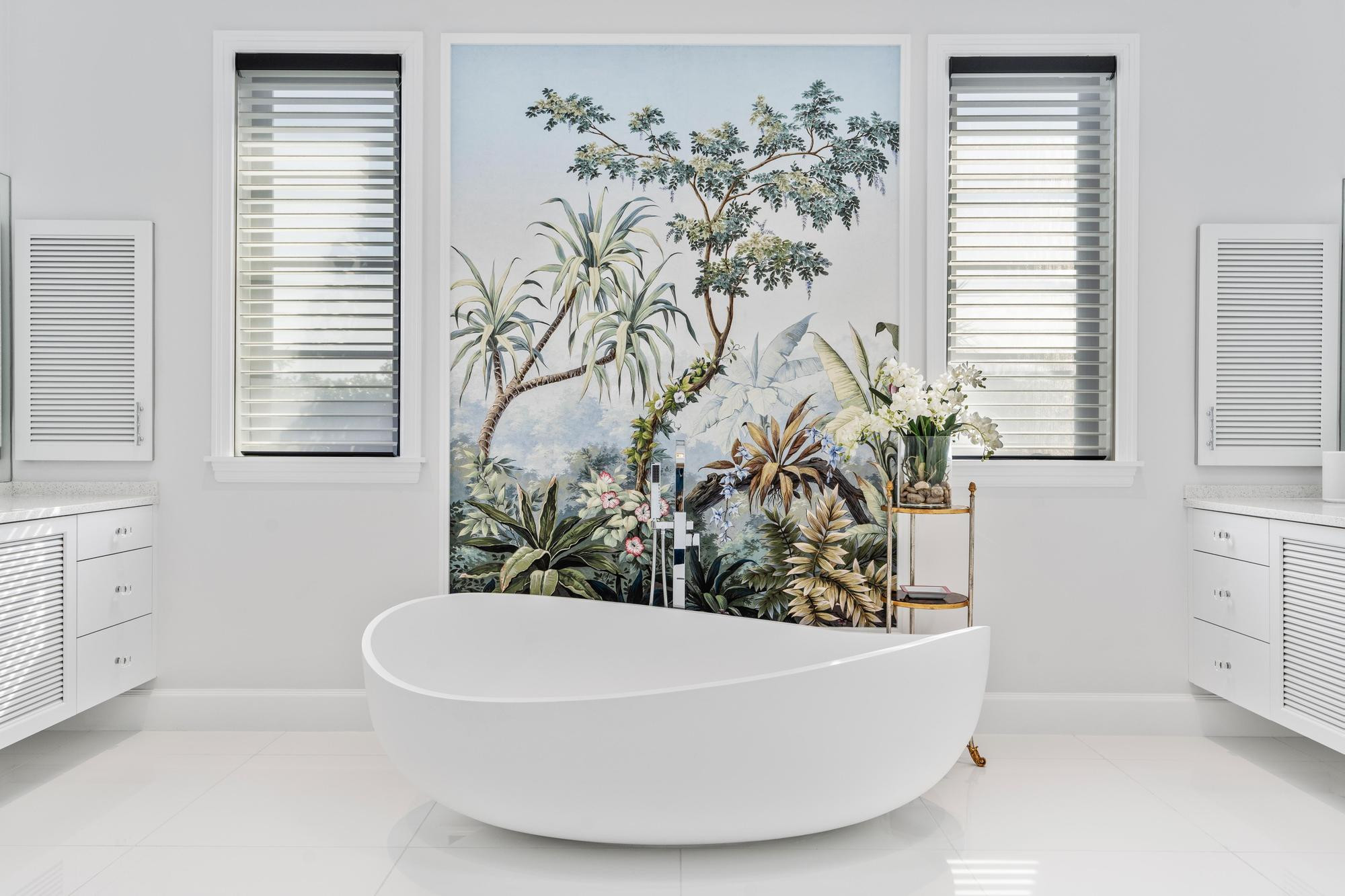 Freestanding oval soaking tub with a floor-mounted chrome filler standing in front of a hand-painted tropical palm and foliage mural, framed by two windows with layered sheer shades, white louvered cabinetry and a brass side table with white orchids.