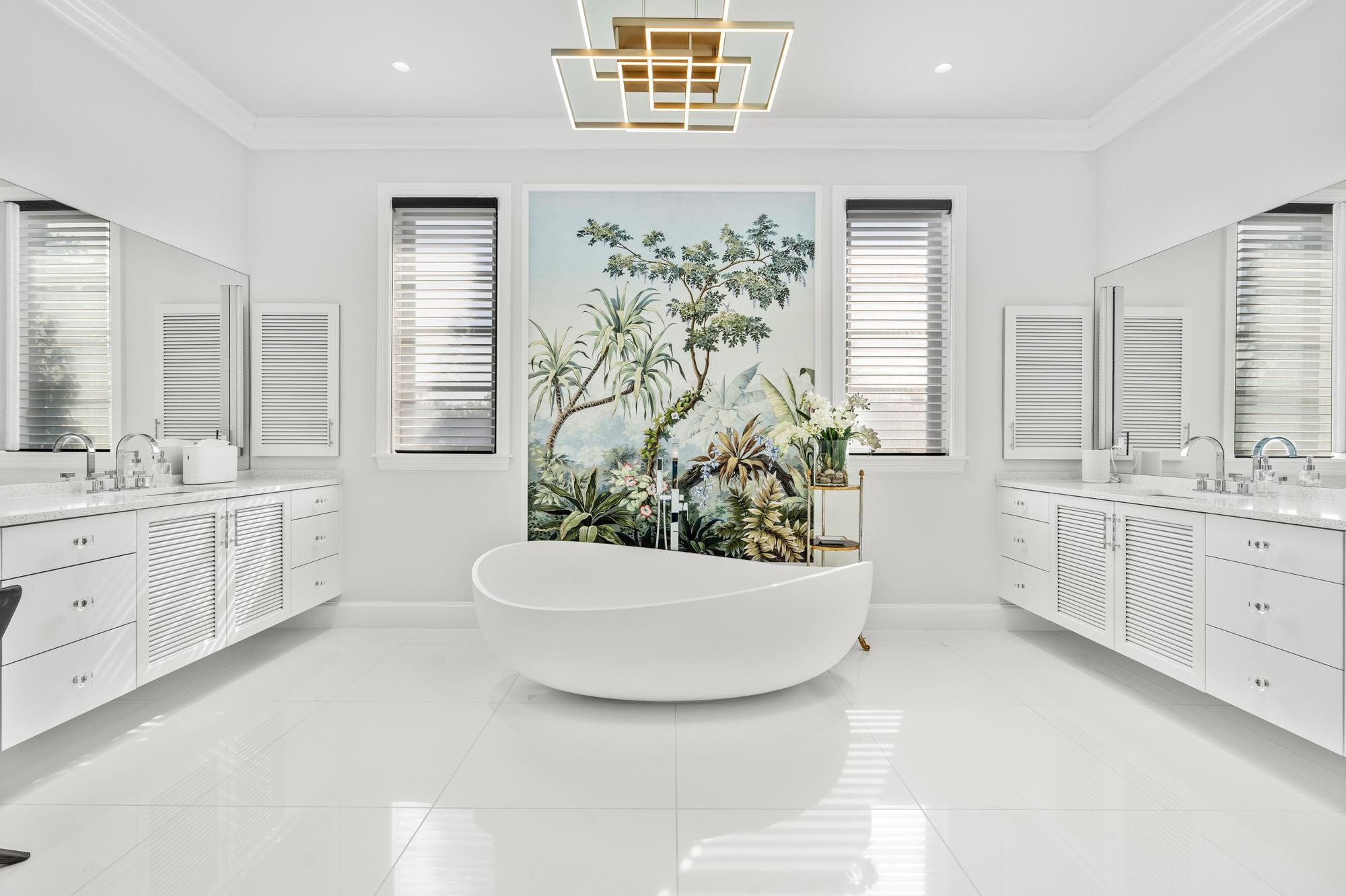 Symmetrical primary bathroom with twin white louvered vanities, speckled quartz countertops and chrome fittings, a freestanding oval soaking tub centered on a hand-painted tropical mural between two windows, a brass geometric ceiling fixture and glossy white large-format floor tile.