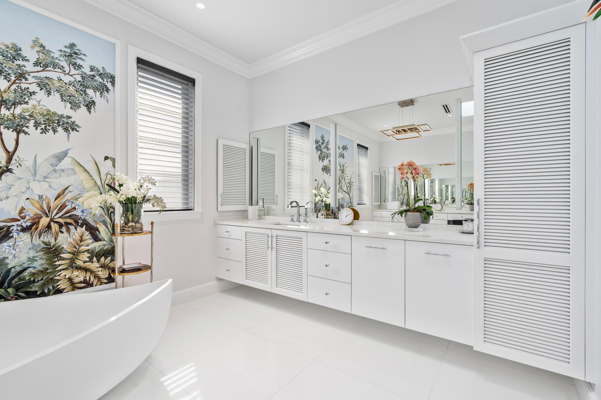 Primary bathroom vanity wall with a full-width mirror, white louvered and slab cabinetry with chrome hardware, a speckled quartz countertop with undermount sinks, orchids and a tall louvered linen tower, with the tropical mural and the soaking tub at left.