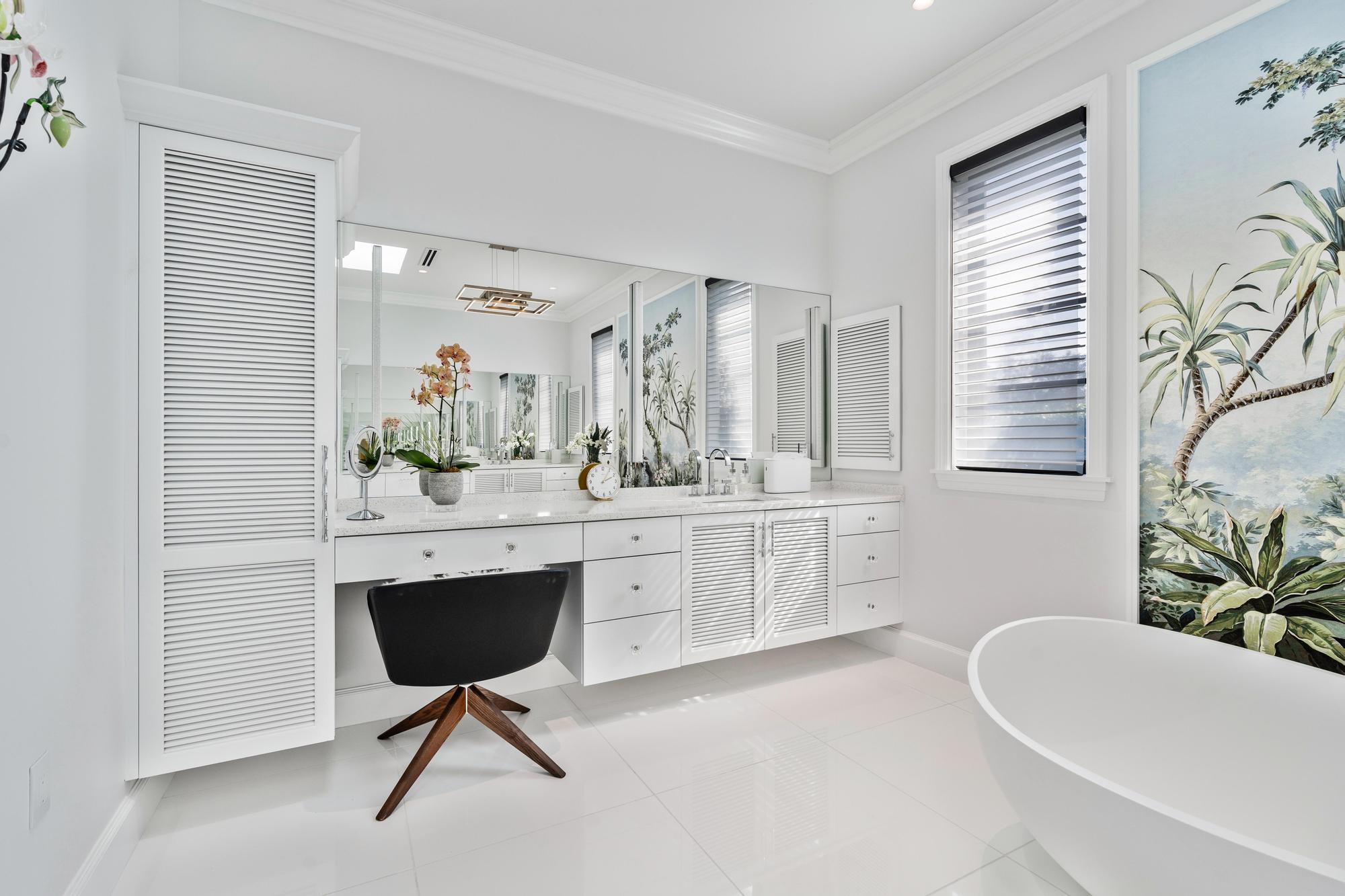 Makeup vanity niche with a white quartz counter, acrylic knobs, a black upholstered swivel chair on a walnut base, a full-width mirror and a tall white louvered linen tower, with the tropical mural and the freestanding tub on the right.