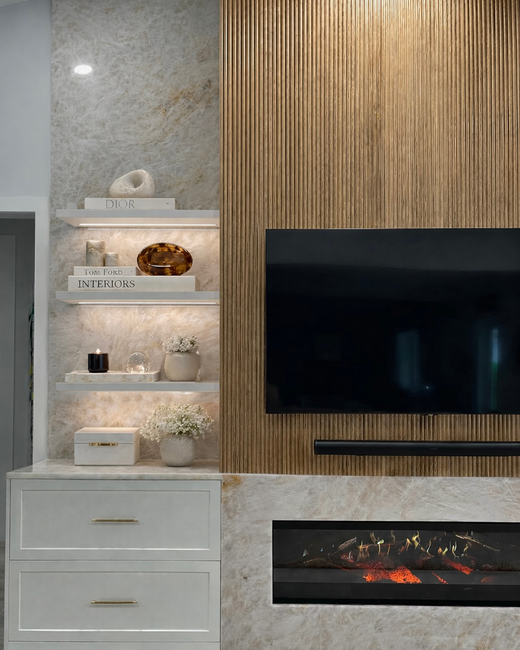 Custom media wall in fluted white oak with a wall-mounted television and soundbar, backlit floating shelves against a translucent quartzite slab, white cabinetry with brass pulls and a linear fireplace in a stone surround
