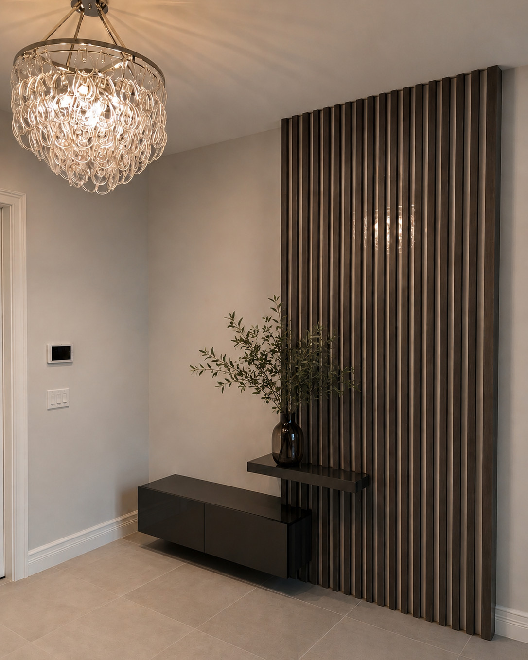 Entry foyer with a floor-to-ceiling dark wood slat wall, a floating black lacquer console and shelf, an olive branch arrangement in a smoked glass vase, and a glass-loop chandelier over large-format beige floor tile.