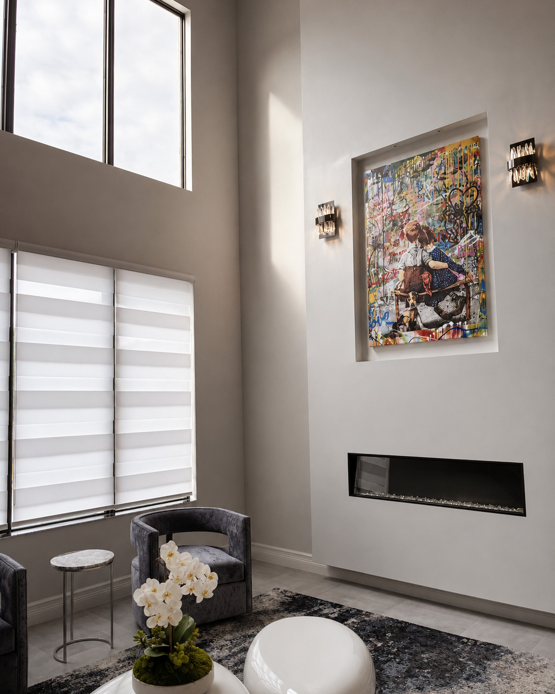 Double-height living room with a recessed wall niche holding a large graffiti-style canvas, crystal sconces on either side, a linear gas fireplace, roller shades and gray velvet swivel chairs.