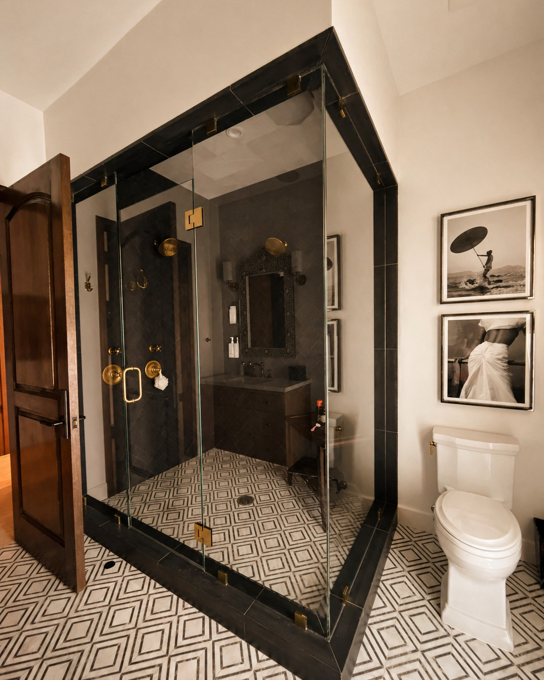 Bathroom with a glass shower enclosure framed in black stone with brass clips and hardware, dark herringbone tile inside, a black and white geometric marble floor, a white toilet and framed black and white photographs.