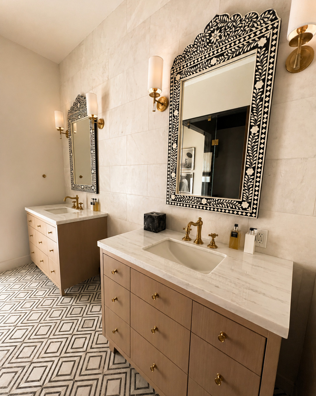 Bathroom with two oak vanities, marble countertops, bone inlay mirrors, brass cross-handle faucets and sconces, and a black and white geometric marble floor.