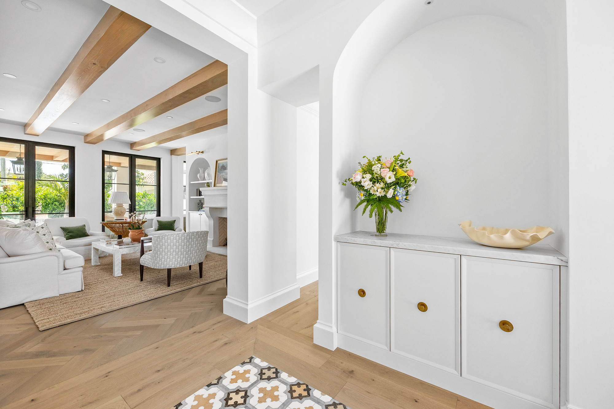 Entry vestibule with an arched niche holding white cabinetry with brass knobs and a marble top, opening onto a beamed living room.
