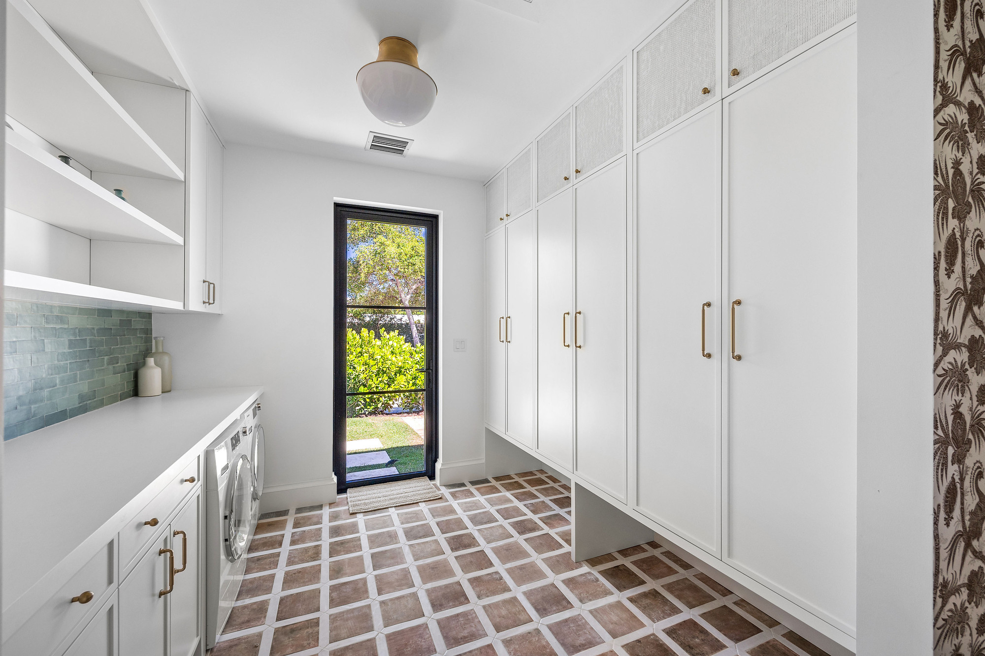 Galley laundry and mudroom with floor-to-ceiling white cabinetry and brass pulls, green glazed tile backsplash and terracotta patterned floor tile.