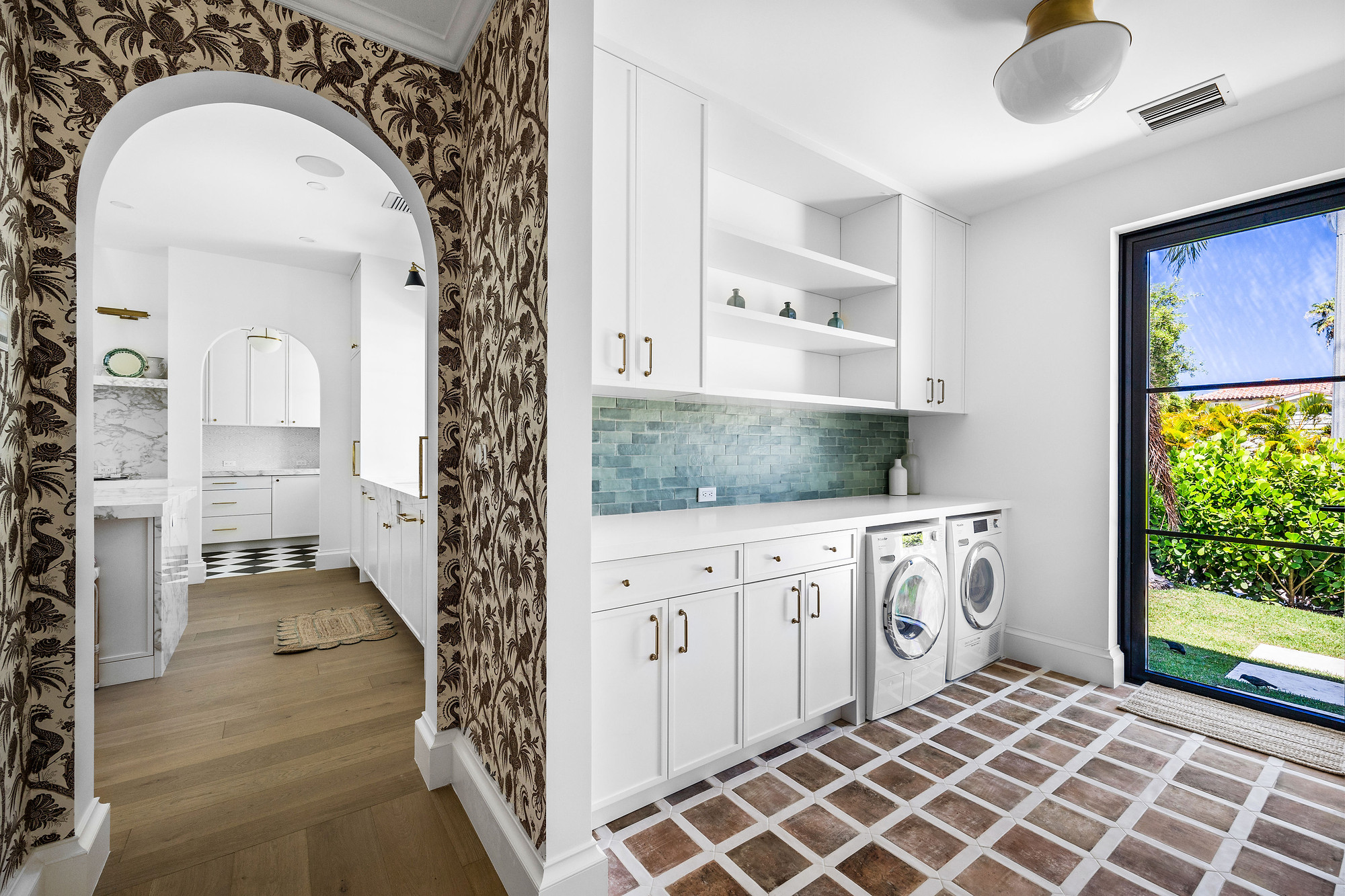 Laundry room with white shaker cabinetry, green tile backsplash and terracotta floor tile, beside a botanical wallpapered archway leading to a butler's area.