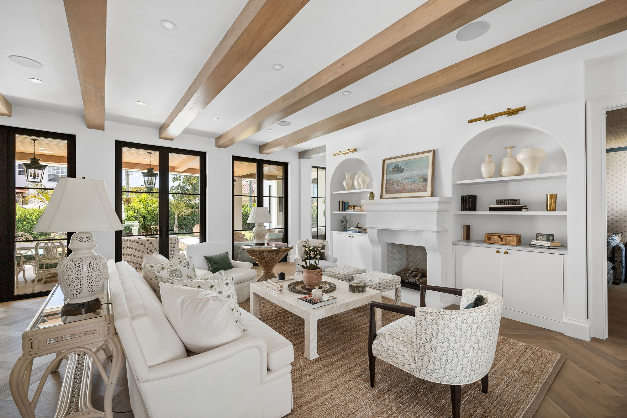 Living room with exposed oak beams, a cast stone mantel set between arched niches with white cabinetry, and black steel French doors opening to the garden.