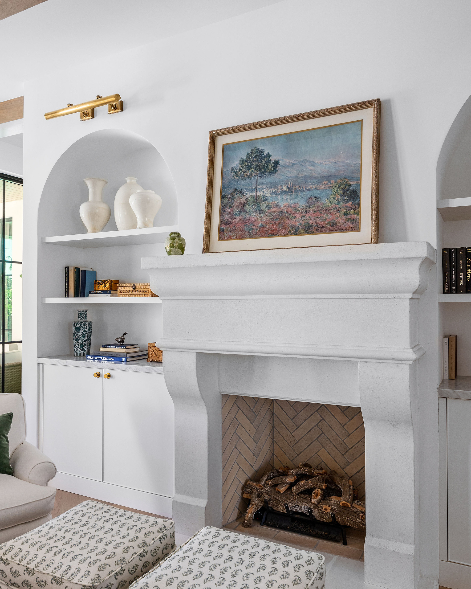 Cast stone fireplace with a herringbone brick firebox, framed by arched plaster niches with open shelving, a brass picture light and white base cabinets.