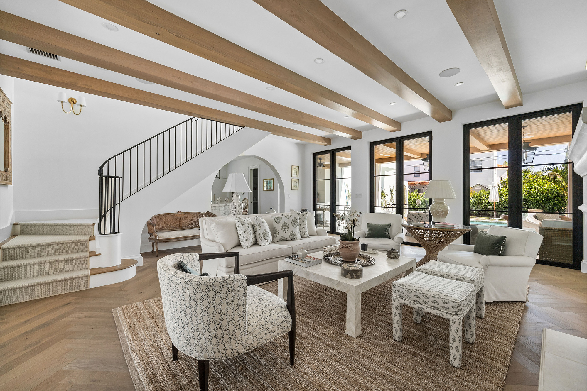 Living room with a beamed ceiling, a curved staircase with black iron railing, and black steel doors opening onto a covered terrace and pool.