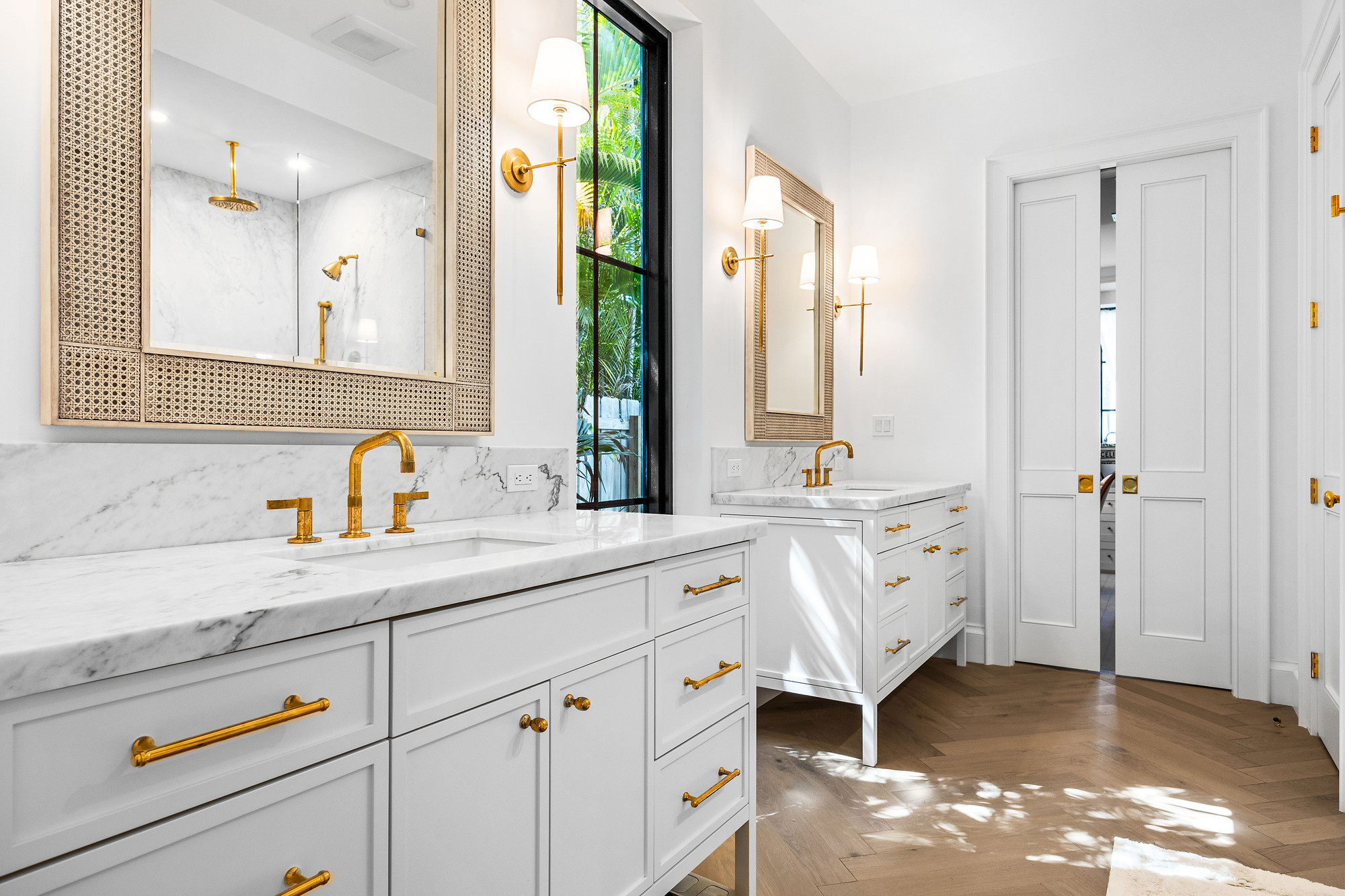 Primary bathroom with two white vanities, marble countertops and backsplash, unlacquered brass fixtures, caned mirrors and herringbone wood flooring.