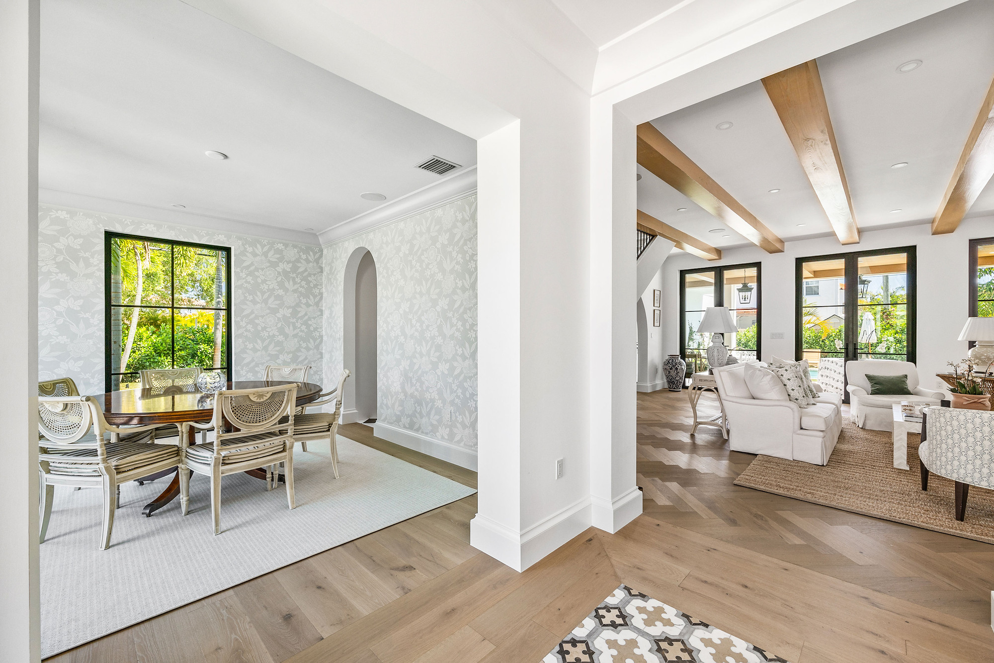 Open plan view of a dining room with soft floral wallpaper and black-framed windows, next to a beamed living room, over wide-plank oak flooring.