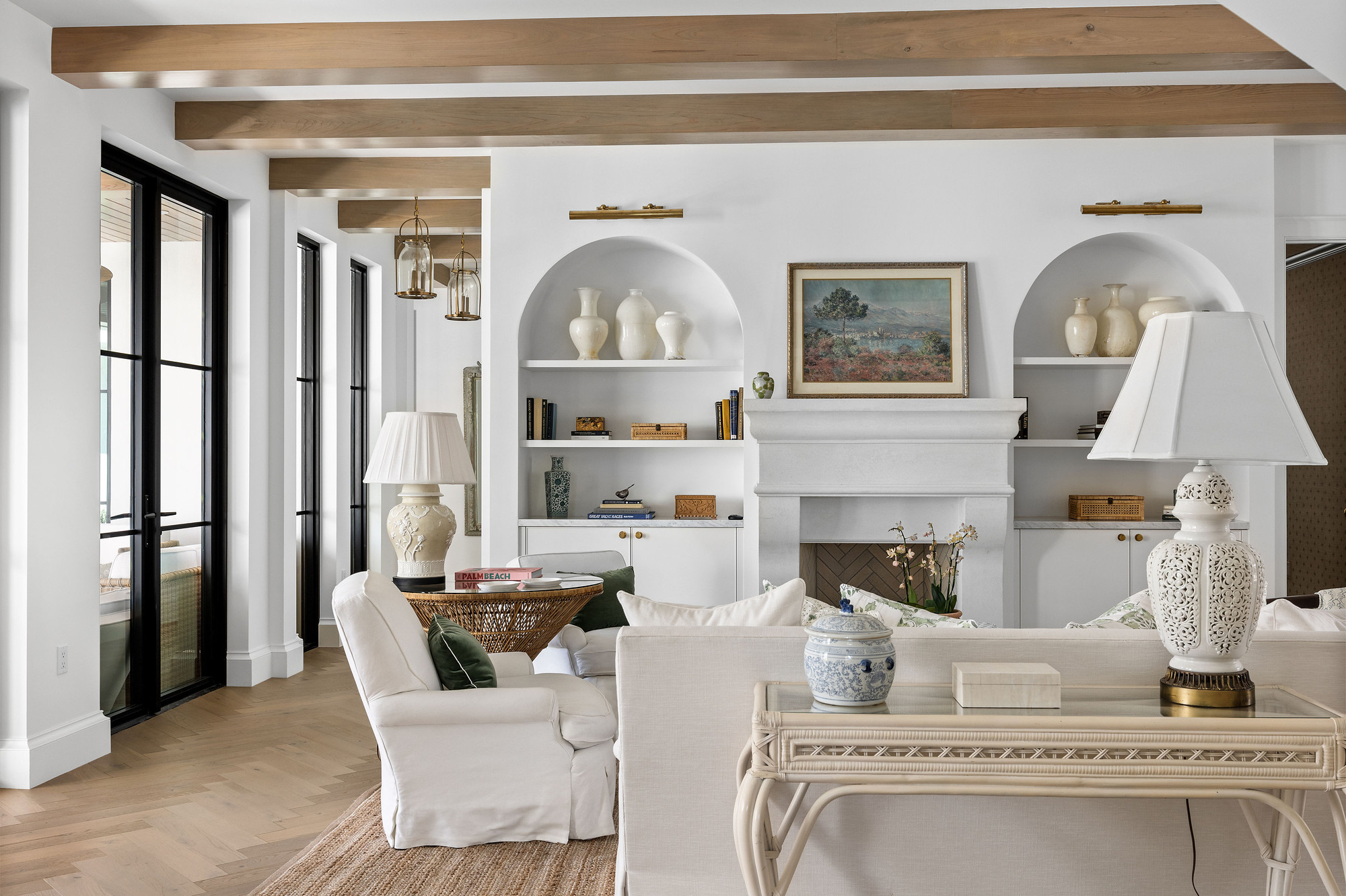Fireplace wall with arched niches, open shelving and brass picture lights above white cabinetry, under light oak ceiling beams.