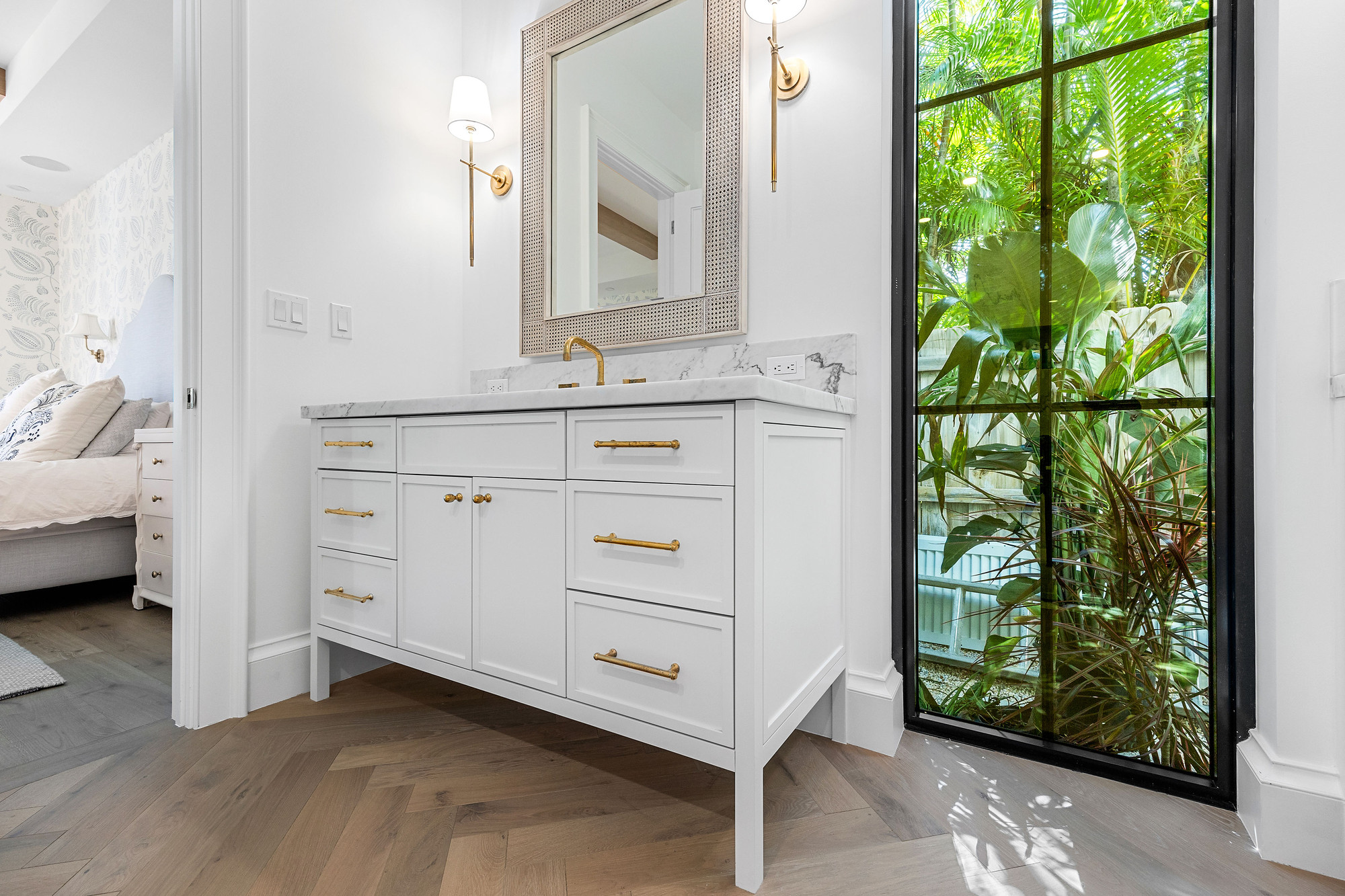 Built-in vanity with marble top and brass pulls beside a tall black steel window, opening to a bedroom with herringbone wood flooring.