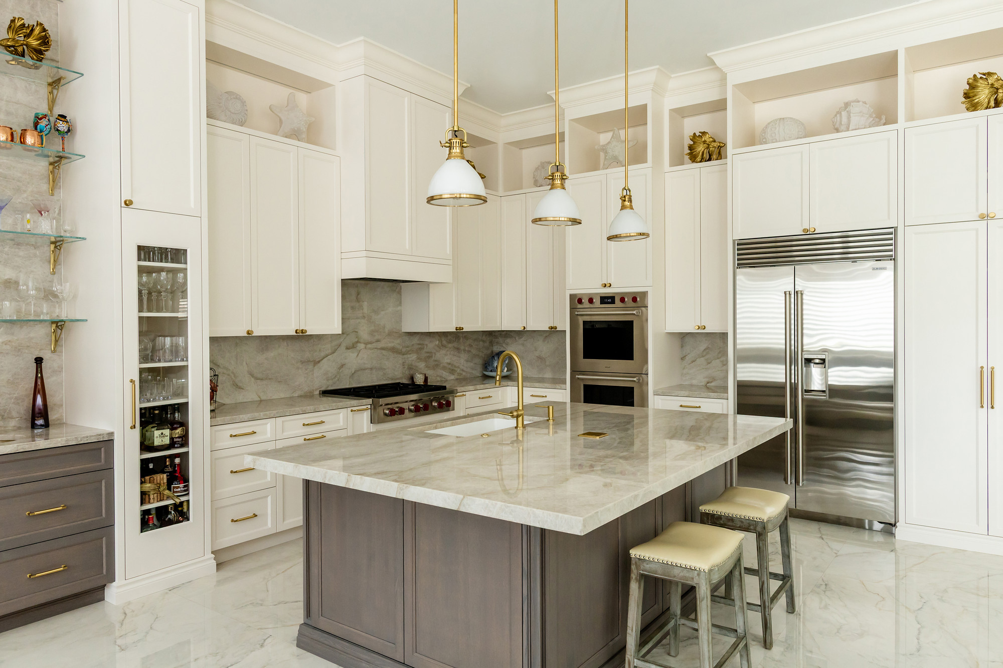 Wide view of an ivory kitchen with a large grey-stained island topped in quartzite, three brass and white dome pendants, professional stainless range, double wall ovens, built-in refrigerator and polished marble flooring.