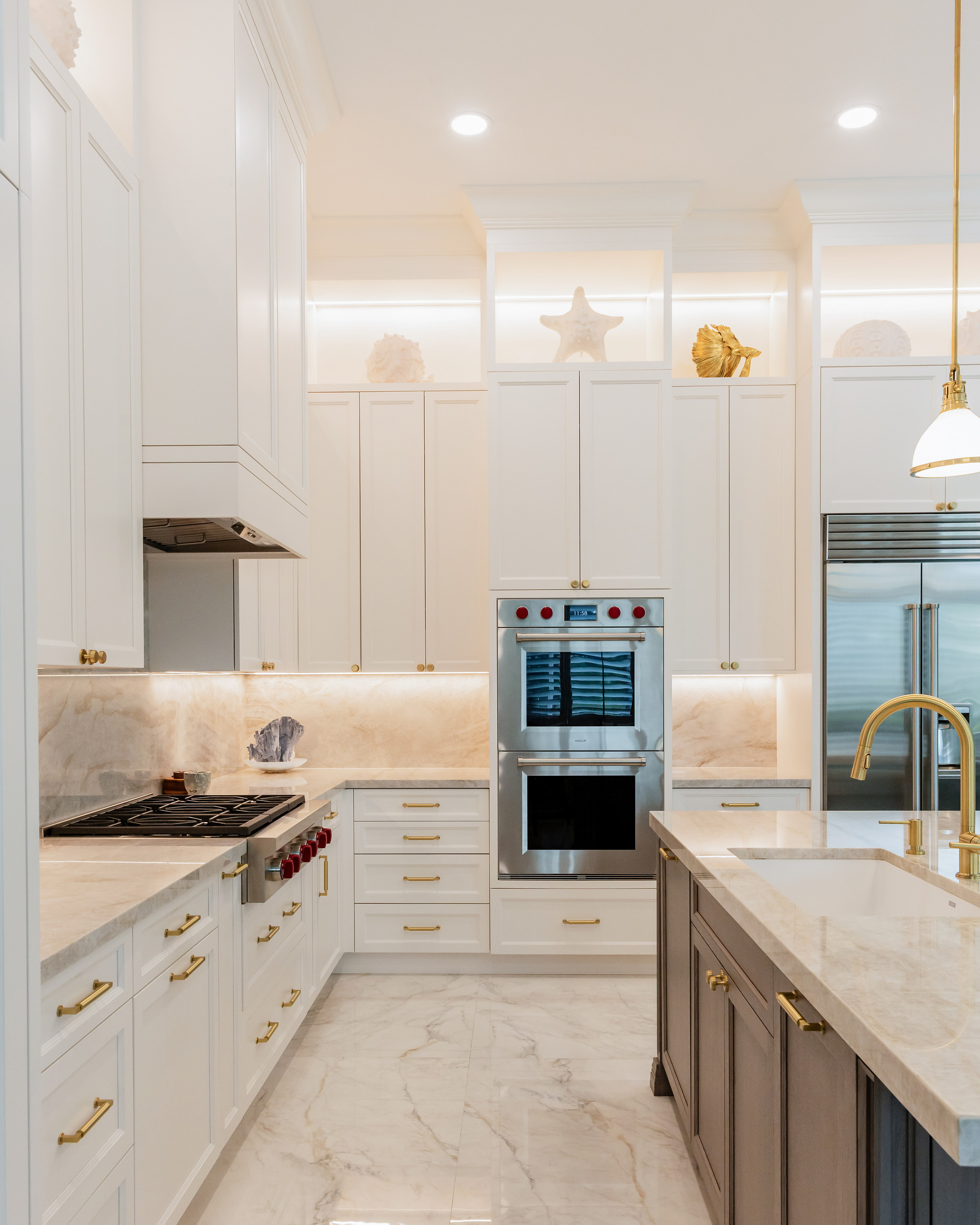 Ivory kitchen corridor view with under-cabinet lighting washing a quartzite backsplash, stainless double wall ovens, gas cooktop, brass pulls, brass faucet over the island sink and polished marble floor.