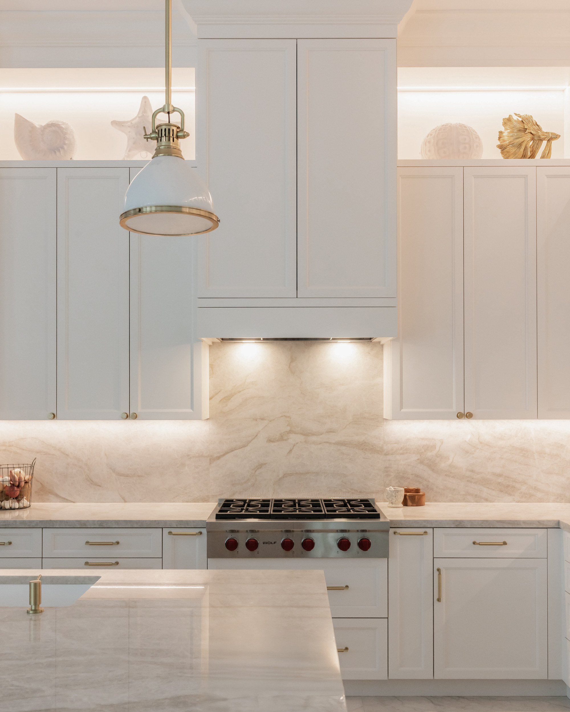 Range wall with a paneled ivory hood, backlit quartzite backsplash, professional stainless cooktop with red knobs, brass hardware and an illuminated display niche with coral and shell objects.