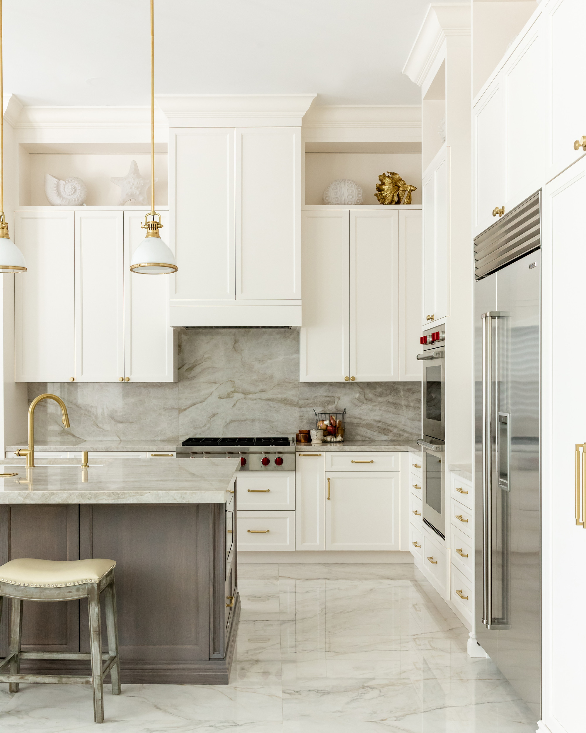 Ivory inset cabinetry with brass pulls, quartzite slab backsplash, professional stainless range, grey-stained island with stone top and a weathered wood stool over polished marble flooring.