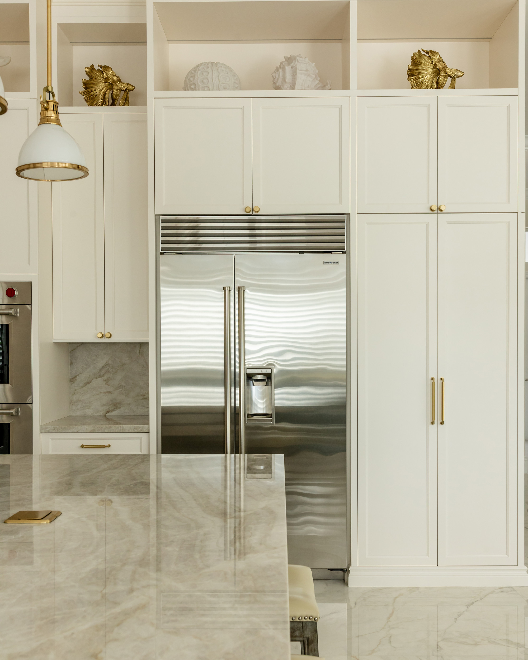 Elevation of ivory floor-to-ceiling cabinetry with open display niches, gilded coral sculptures, a built-in stainless refrigerator and a pendant light above a quartzite island countertop.
