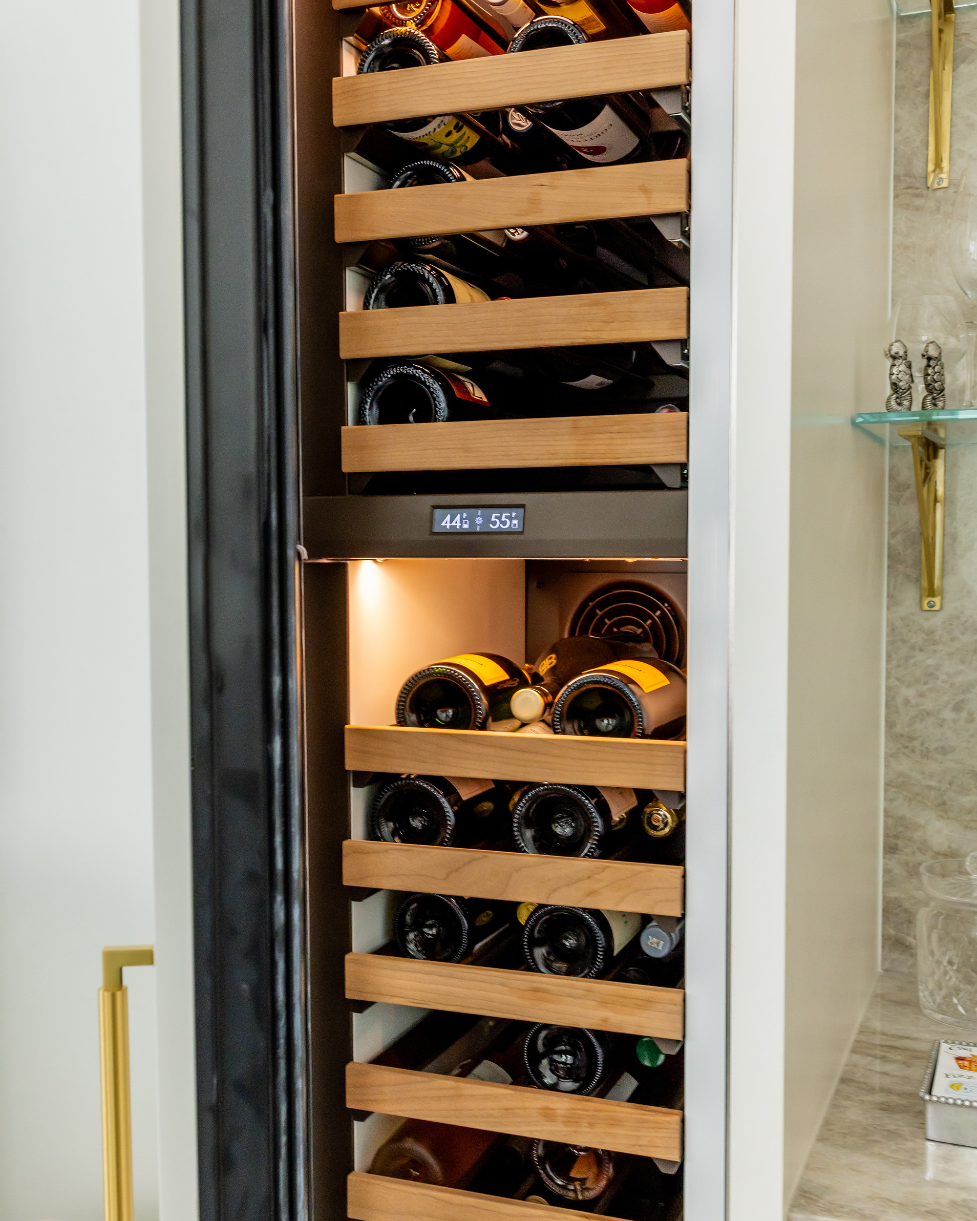 Open dual-zone wine column with wooden pull-out racks, lit interior and a digital temperature display, framed by ivory cabinetry and brass hardware.