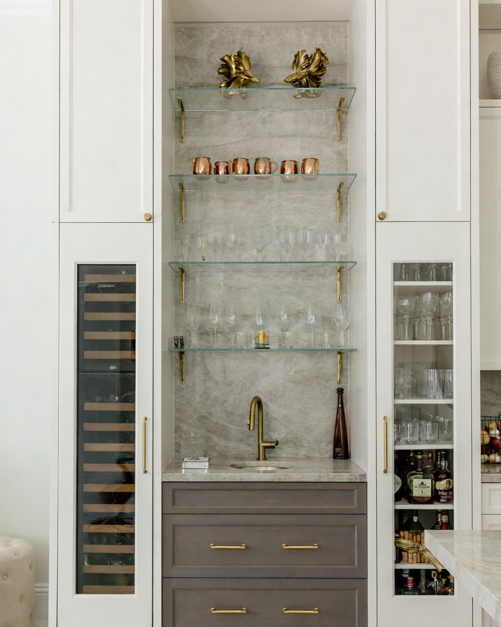 Wet bar alcove with a quartzite slab backsplash, four backlit glass shelves holding copper mugs and crystal stemware, a gray-brown wood cabinet with brass bar pulls, flanked by a built-in wine refrigerator and a glass-front liquor cabinet.