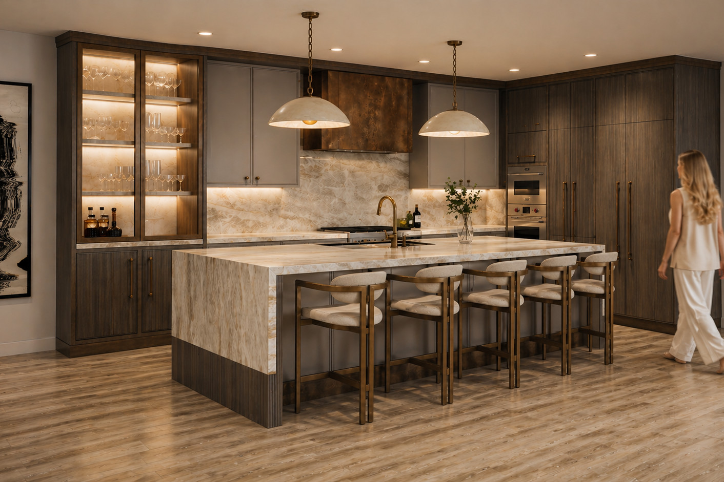 Wide kitchen view with a pale quartzite waterfall island on a reeded wood plinth, five brass-framed cream counter stools, dark oak tall cabinetry, a bronze hood and a lit glass bar cabinet on a light oak floor.