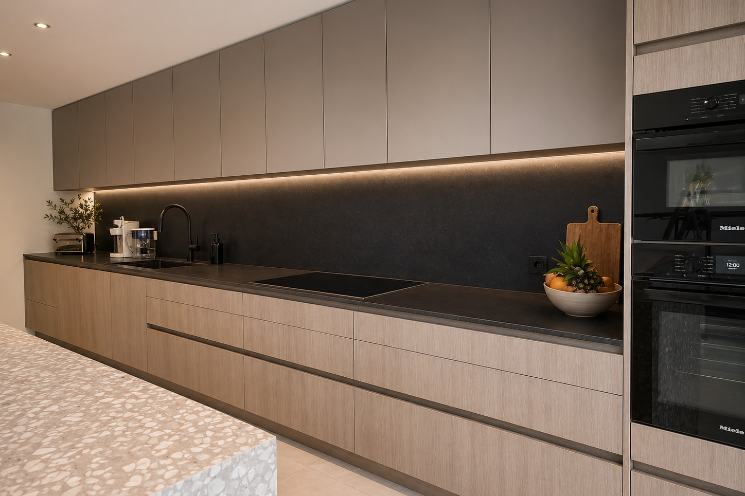 Handleless kitchen with pale oak base cabinets, matte taupe upper cabinets, dark stone backsplash and countertop, integrated LED strip lighting, black built-in ovens in a tall oak unit and a terrazzo island in the foreground.