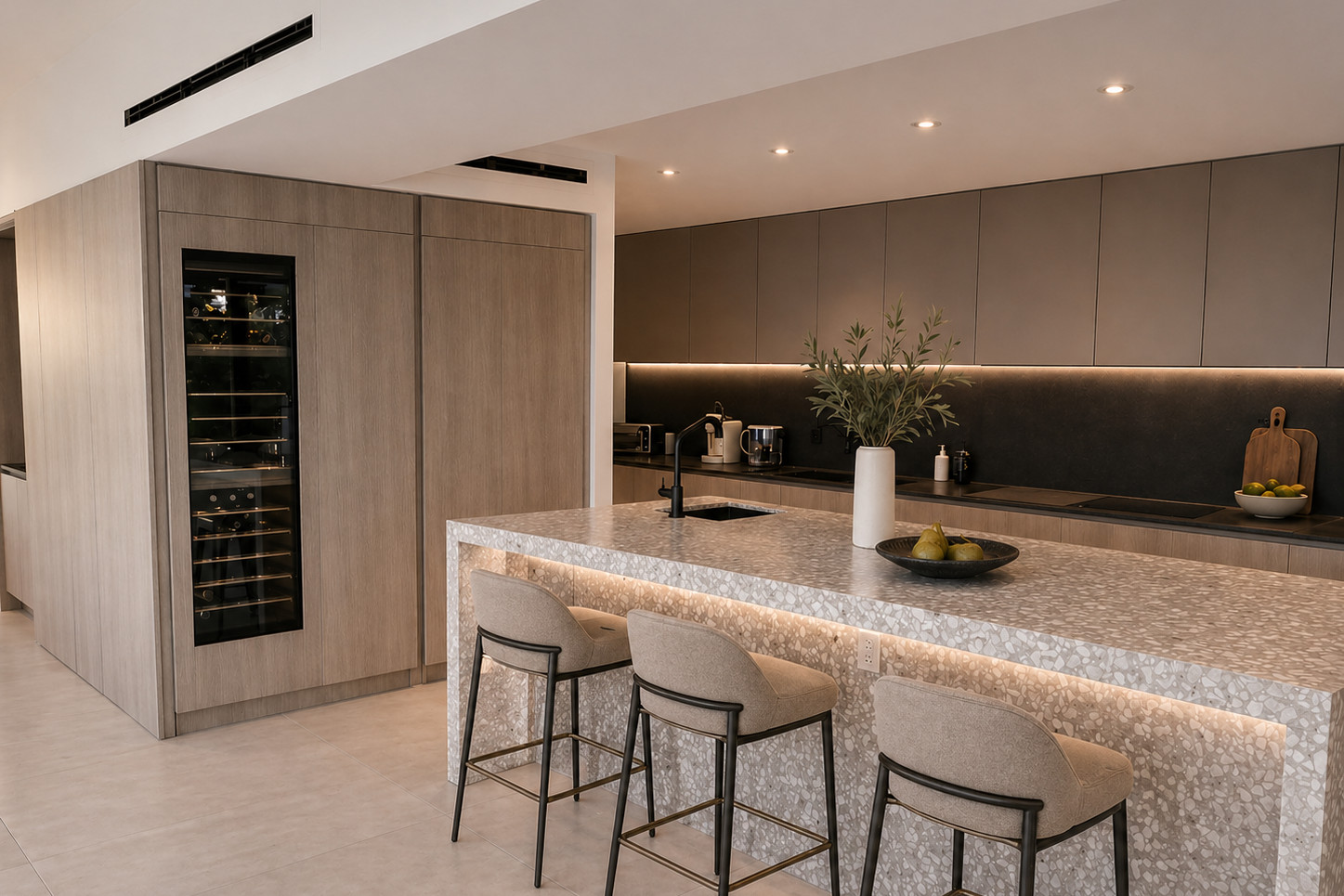 Open kitchen with a grey terrazzo waterfall island and three upholstered bar stools, a backlit recess under the counter, pale oak tall cabinetry with an integrated wine column, matte taupe wall cabinets and a dark stone backsplash.